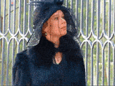 a woman wearing a black hat and fur coat is standing in front of a fence