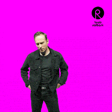 a man in a black denim jacket stands in front of a pink background with a logo for teatr rozbark on it