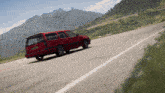 a red car is driving down a road with mountains in the back