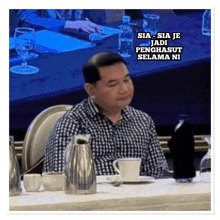 a man in a plaid shirt sits at a table with a caption that says sia-sia je jadi penghast selama ni