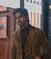 a man wearing a brown jacket and a black turtleneck is standing in a room .