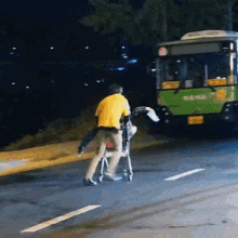 a man in a yellow shirt is pushing a child in a walker down a street in front of a green bus