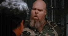 a man with a beard and a camouflage shirt is talking to another man in a jail cell .