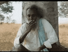 an elderly man with a beard is smoking a cigarette while sitting under a tree .