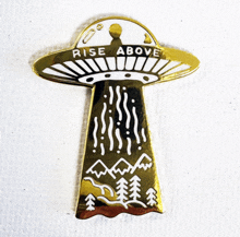 a gold pin with a flying saucer and the words rise above
