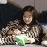 a woman in a striped shirt is sitting on a couch holding a green bag of chips .