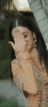 a woman in a dress is covering her face with her hand while standing next to a tree .