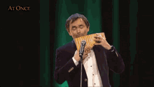 a man singing into a microphone while holding a flute in front of a green curtain that says at once