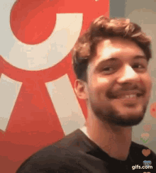 a young man with a beard is smiling in front of a red and white sign .