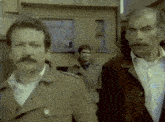 a man with a mustache is standing next to another man in a trench coat .