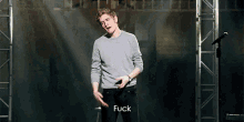 a man in a grey sweater is standing in front of a microphone with the word fuck above him