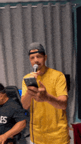 a man in a yellow shirt is singing into a microphone while holding a cellphone