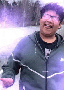 a young man wearing glasses and a nike jacket is laughing and pointing at the camera .
