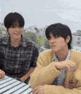 two young men are sitting at a table and one of them is wearing a yellow sweater with a t on it