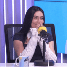 a woman covering her mouth while sitting in front of a microphone and a mug that says ' a ' on it