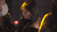a woman wearing a black off the shoulder top stands in front of a microphone with a red light behind her