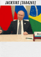 a man in a suit and tie is sitting at a table with flags in the background and a sign that says ru on it
