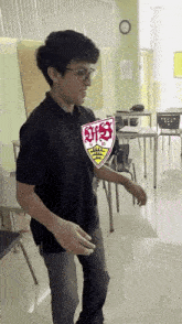 a young man is standing in a classroom wearing a shirt with a shield on his chest .