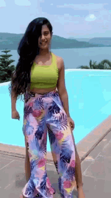a woman in a green top and floral pants is standing next to a pool .