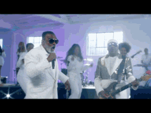 a man in a white suit is singing into a microphone while another man plays a guitar