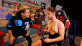 a man without a shirt is talking to another man in a gaming chair with the letter a on it