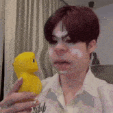 a person holding a yellow rubber duck with white paint on their face and a shirt that says " why " on it