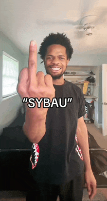 a man wearing a black shirt that says " sybau " shows his middle finger