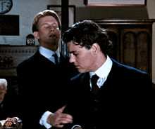 two men in suits and ties are talking in a room with a clock on the wall