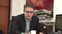 a man in a suit and glasses sits at a desk holding a cup of coffee