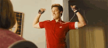 a man in a red shirt is flexing his muscles while wearing headphones and holding a remote control .