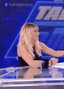 a woman is sitting at a table with a mug in front of a large screen .