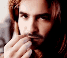 a man with long hair and a beard is holding his hand to his mouth .