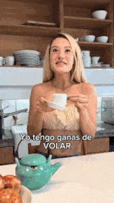 a woman in a kitchen holding a cup and saucer with yo tengo ganas de volar above her