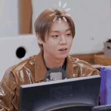 a young man in a brown jacket is sitting in front of a computer monitor with a microphone in his hand