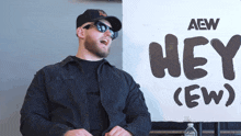 a man wearing sunglasses and a hat stands in front of a sign that says hey ( ew )