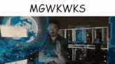 a man is sitting in front of a screen with the words mgwkwks written above him .