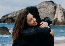 a woman in a black jacket is hugging a man in front of the ocean