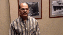 a bald man wearing glasses and a striped shirt stands in front of two framed pictures on the wall