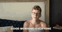 a shirtless man with glasses is sitting on a bed and saying good morning everyone .