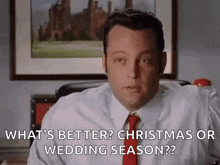 a man in a suit and tie is sitting at a desk in front of a picture and talking about christmas or wedding season .