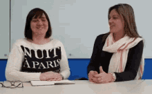 two women are sitting at a table with one wearing a nuit a paris shirt