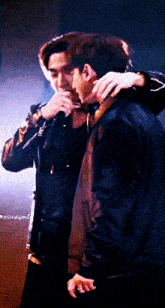 a man in a leather jacket is hugging another man in a dark room