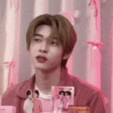 a young man in a pink jacket is sitting in front of a pink wall holding a card .