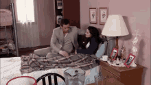 a man in a suit sits next to a woman laying in bed