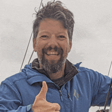 a man with a beard wearing a blue jacket giving a thumbs up sign