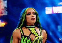 a woman with green hair and blue eyes is standing in front of a blue background with a w logo on it .