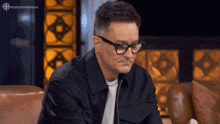 a man wearing glasses and a black jacket is sitting on a couch in front of a sign that says cbcdragonsden