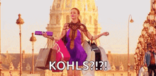 a woman is holding a bunch of shopping bags in front of a building and says `` kohls ? ''