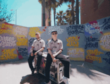 two men sitting on a box in front of a wall with graffiti on it
