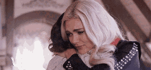 a woman with white hair is hugging another woman with a sad look on her face .